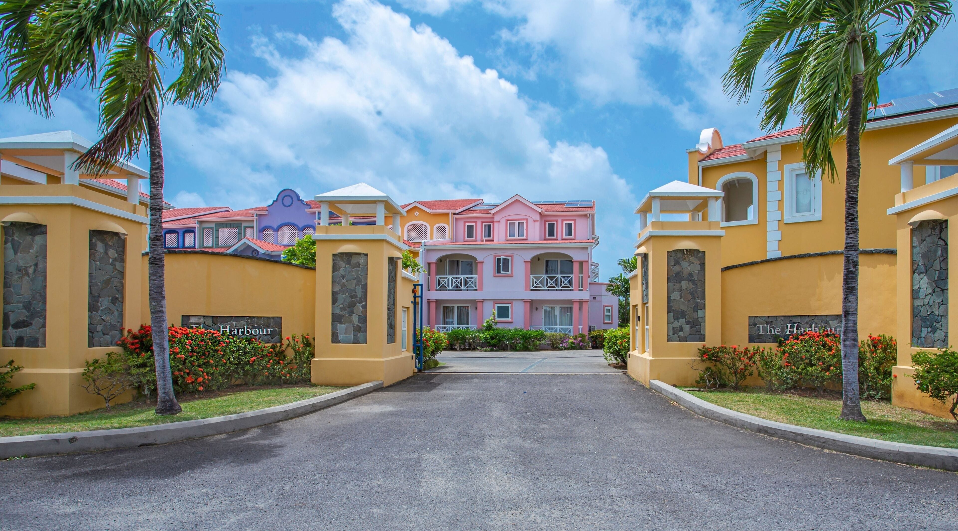 A hotel entrance with a central pathway leading to a pink building with balconies, surrounded by palm trees and red flowers.