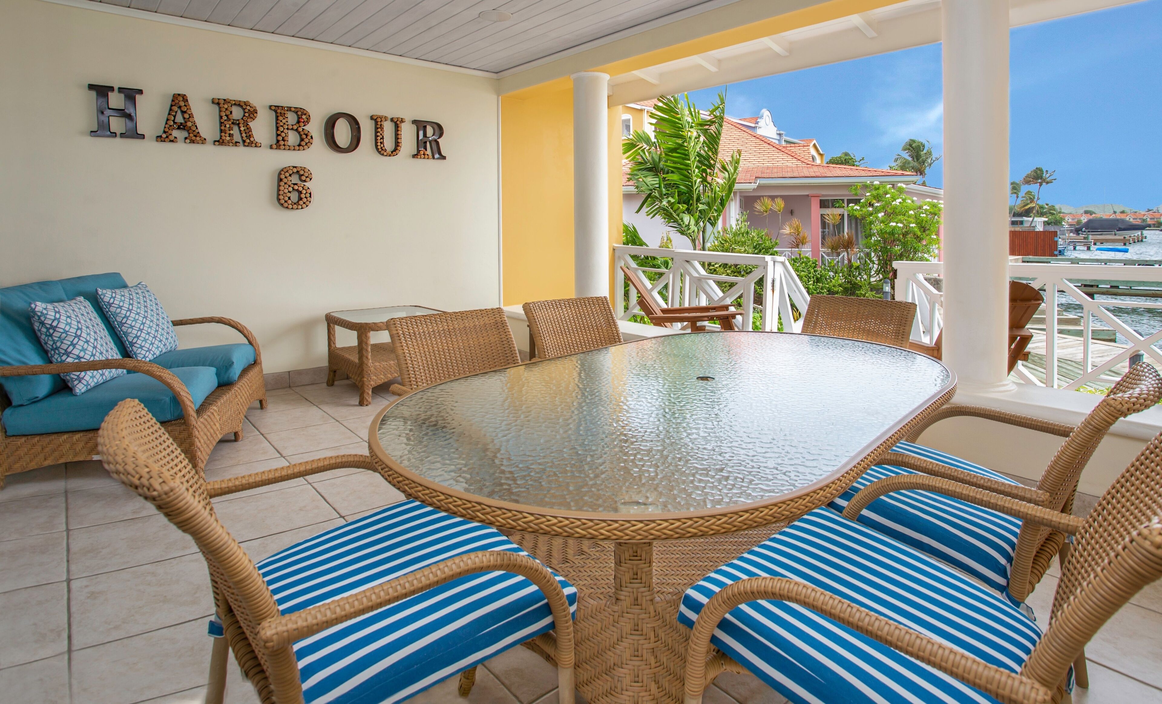 A spacious outdoor dining area with wicker furniture and a glass table, overlooking a scenic harbor.