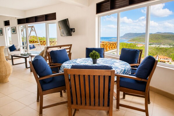 A room with a round table, four chairs, a TV, and a view of the ocean.