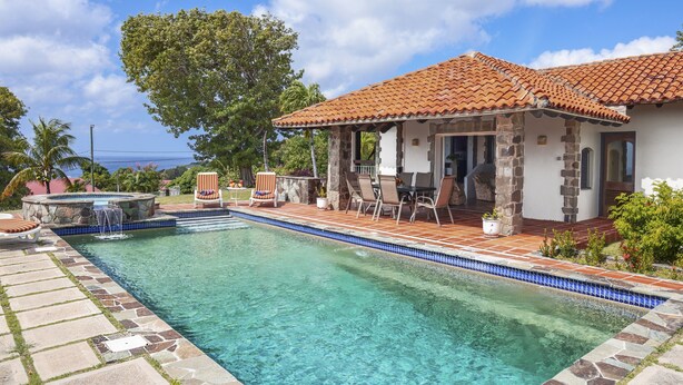 A swimming pool with a tiled deck, a covered patio with outdoor furniture, and a stone building with a tiled roof. - Spanish-style Ocean View Villa Set In Garden - Calypso Court 3 Bedroom Villa by RedAwning (Gros Islet)