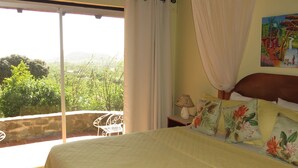A bedroom with a bed, a nightstand, a lamp, a painting, and a balcony with a view of greenery. - Spanish-style Ocean View Villa Set In Garden - Calypso Court 3 Bedroom Villa by RedAwning (Gros Islet)