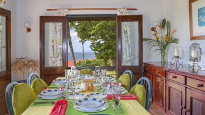 A dining table set with plates, glasses, and cutlery, with a view of the sea through open windows. - Spanish-style Ocean View Villa Set In Garden - Calypso Court 3 Bedroom Villa by RedAwning (Gros Islet)