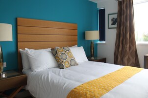 Habitación de hotel con cabecera de madera, ropa de cama blanca, manta amarilla estampada y dos lámparas de noche.