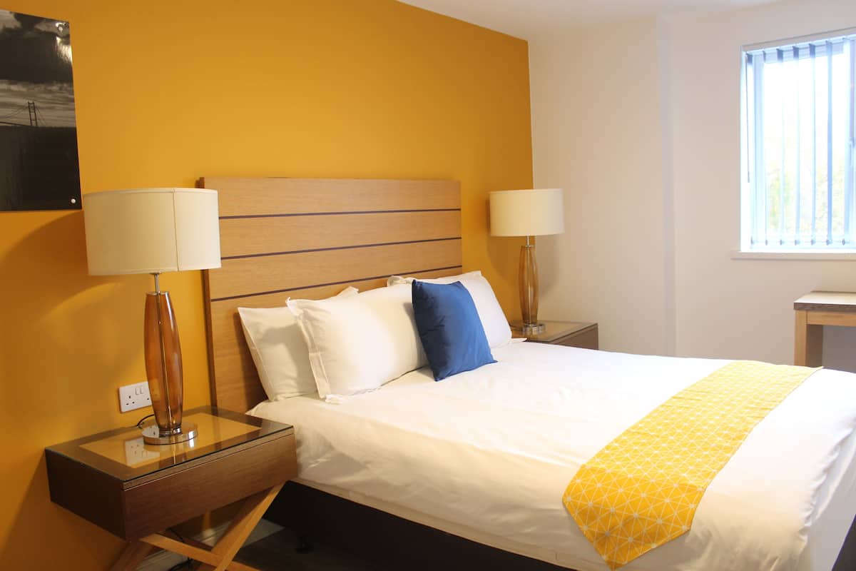 A hotel room with a bed, two bedside lamps, a nightstand, a window, and a wall-mounted picture.