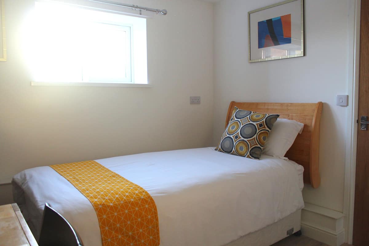 A single bed with a wooden headboard, a patterned pillow, and a white blanket. There is a window with blinds, a framed abstract painting on the wall, and a door with a handle.