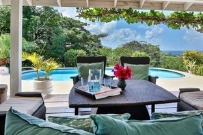 A covered outdoor seating area with wicker furniture, a table with drinks and a book, and a view of a swimming pool and lush greenery.