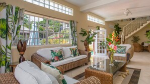 Amplia sala de estar con muebles de mimbre, una mesa de centro de vidrio y ventanas grandes que ofrecen vistas al exterior.