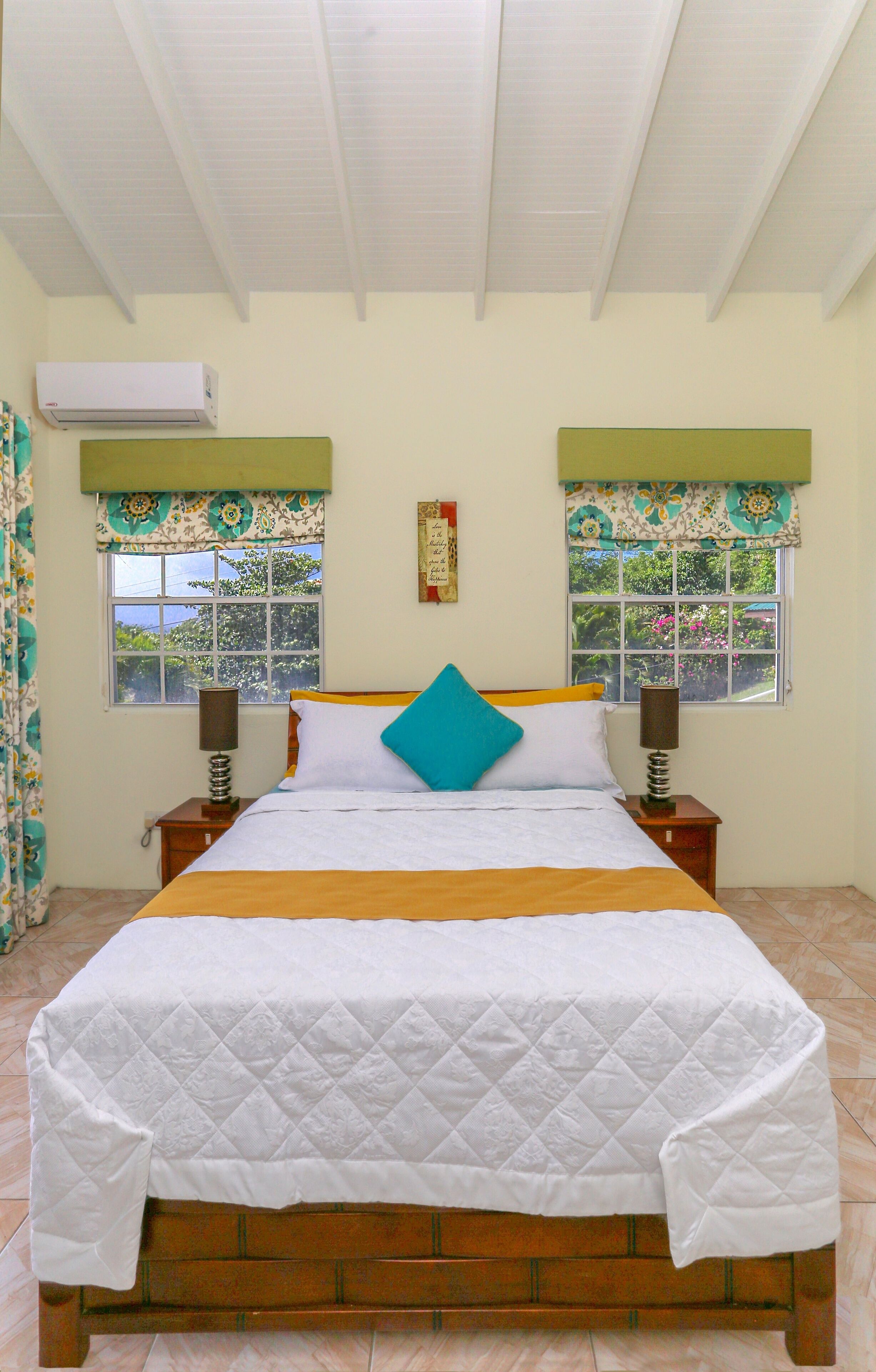 A bedroom with a bed, two bedside tables, a window with curtains, and an air conditioning unit on the wall.