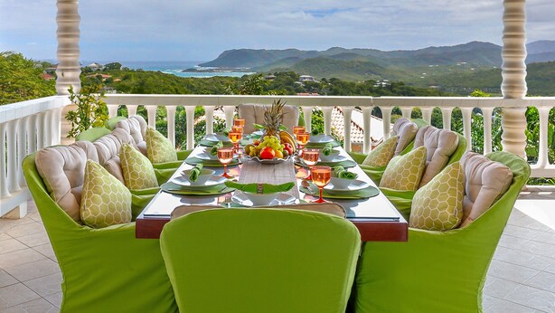 Mesa de comedor preparada para cuatro personas con sillas tapizadas en verde, un frutero, copas de vino y vistas al océano y las colinas.