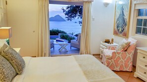 A hotel room with a bed, a small table, a chair, and a view of the sea and mountains.