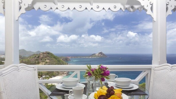 A table with a view of the ocean and hills, a plate of fruit, and a vase with flowers.