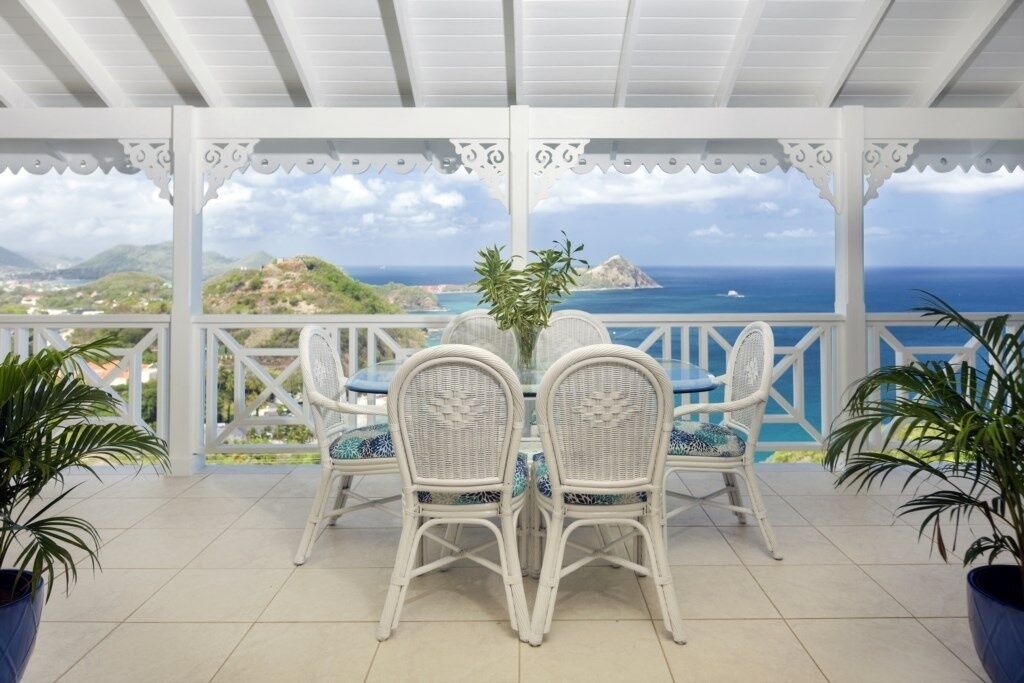 Un patio techado con baranda blanca, una mesa con sillas y vistas al océano y las colinas.