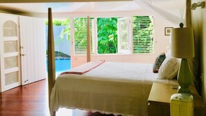 Une chambre avec un lit, un ventilateur de plafond, une lampe de chevet et une vue sur une piscine et la verdure environnante.