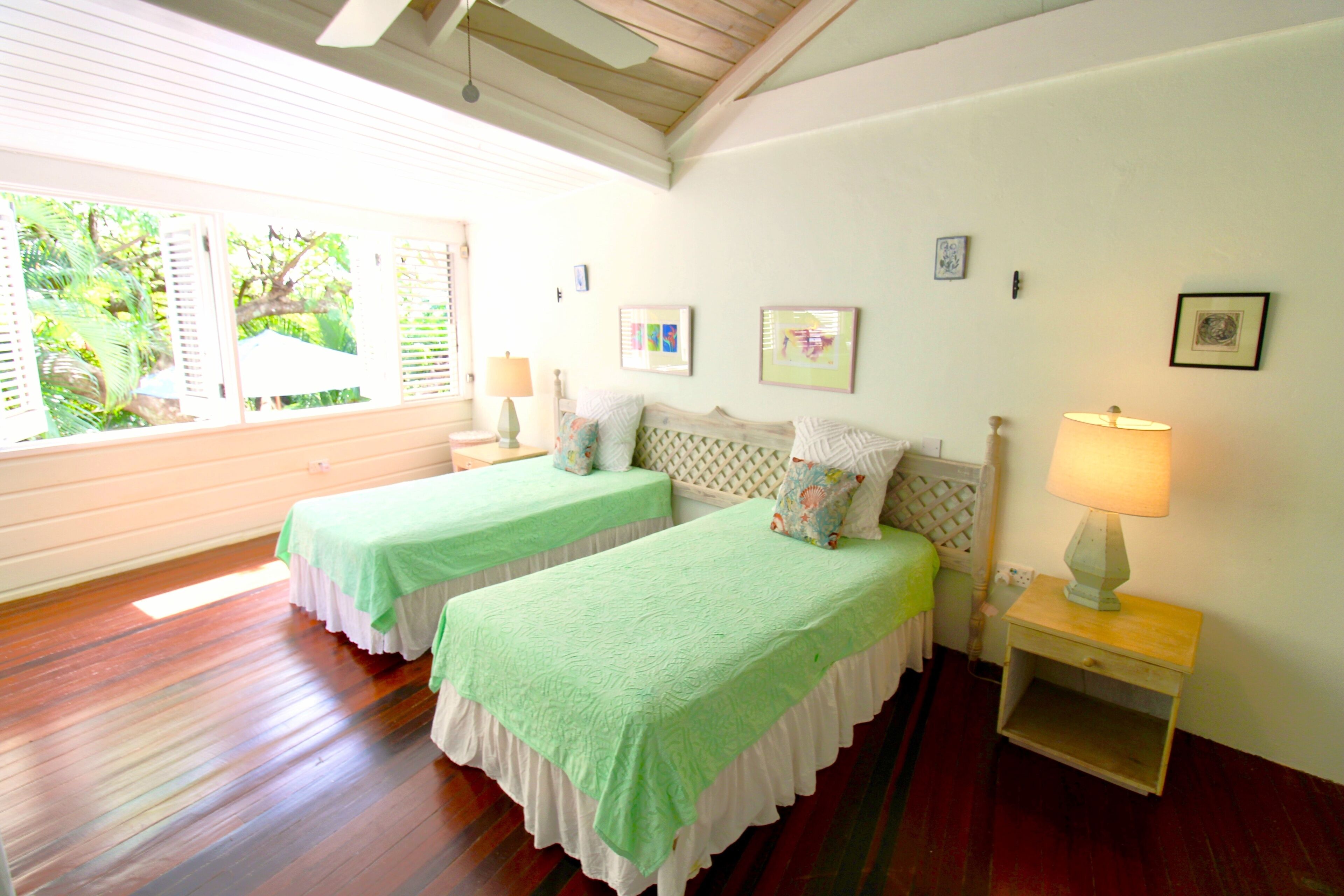 Quarto com duas camas, janela com persianas, mesa de cabeceira com abajur e quadros nas paredes.
