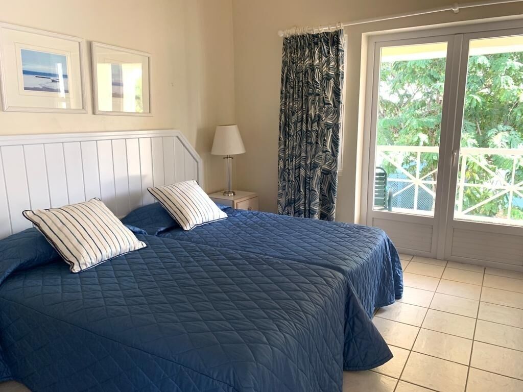 Une chambre avec un grand lit, deux oreillers, une table de chevet avec une lampe, une fenêtre avec des rideaux et une vue sur la verdure à l’extérieur.
