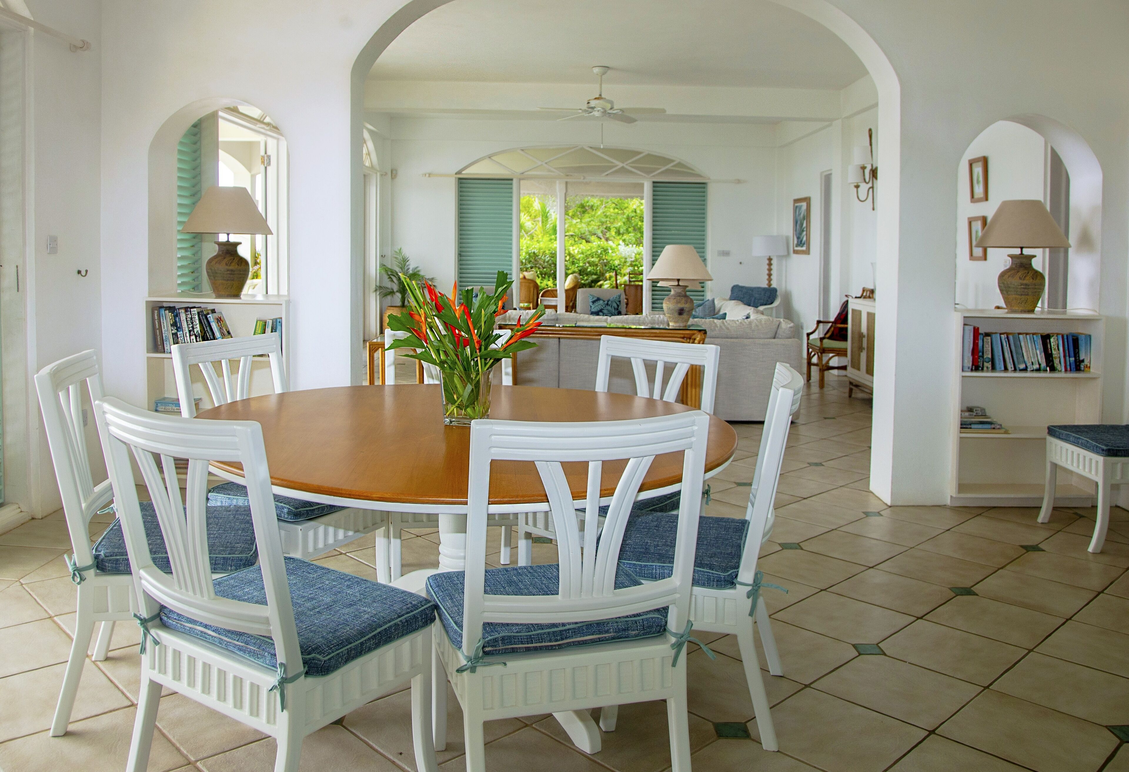 A dining area with a wooden table and white chairs with blue cushions, surrounded by white walls and green shutters.