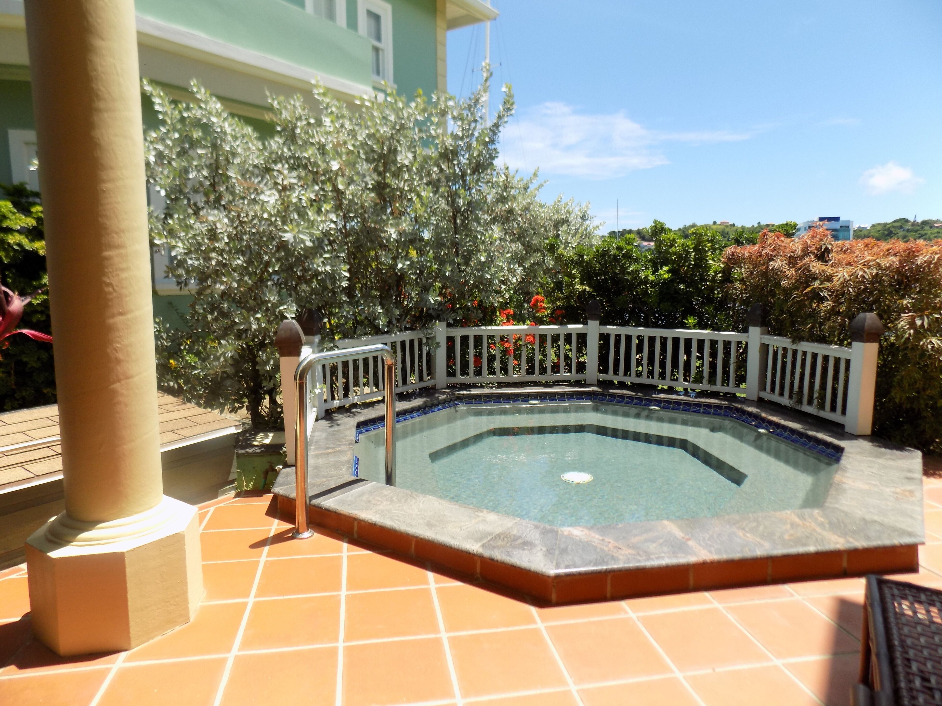 A swimming pool with a white fence, surrounded by a tiled patio and a large tree.
