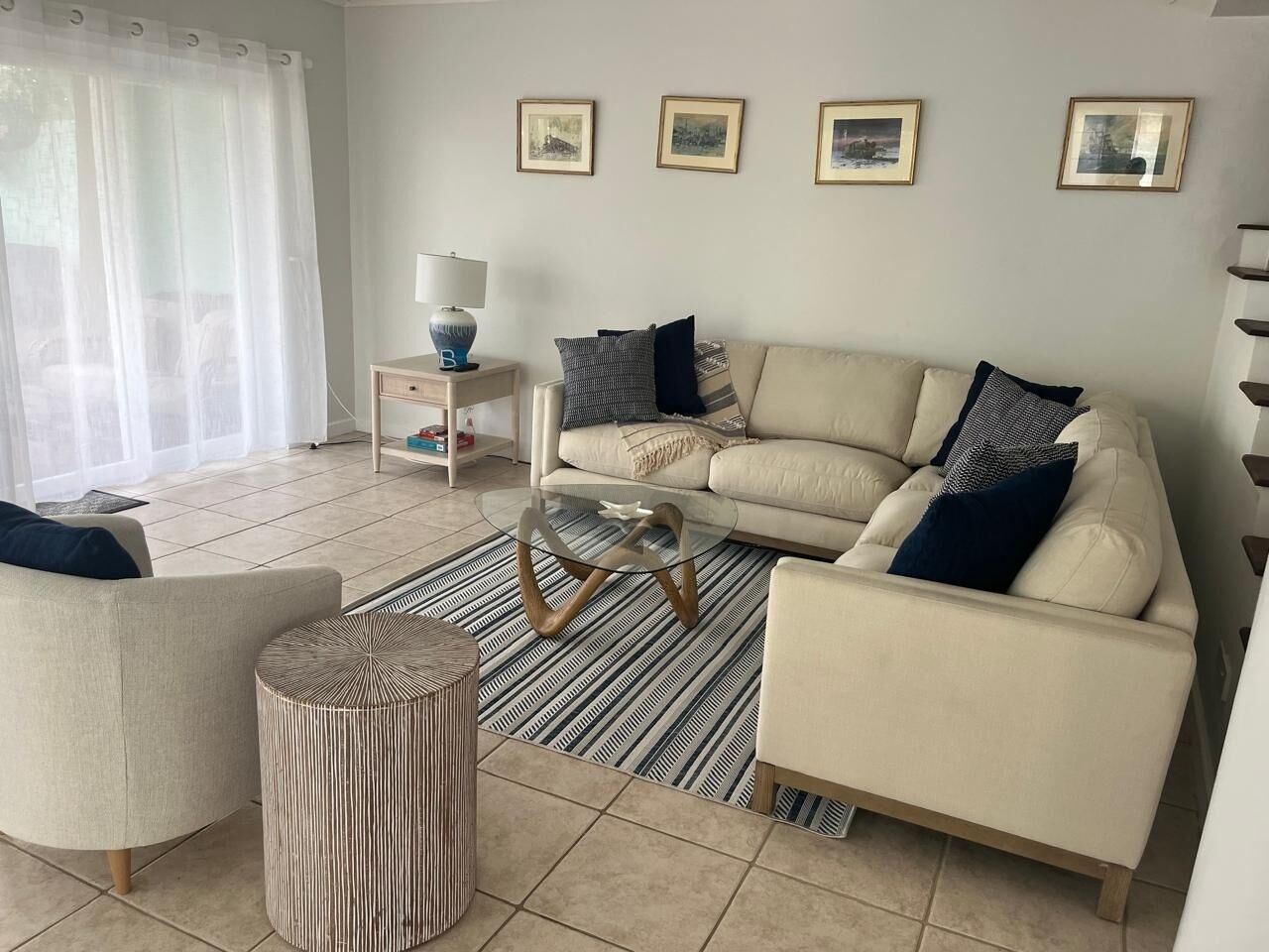 Una sala de estar con un sofá color beige, dos sillones, una mesa de centro de vidrio, un tapete a rayas y una mesita de noche con una lámpara.