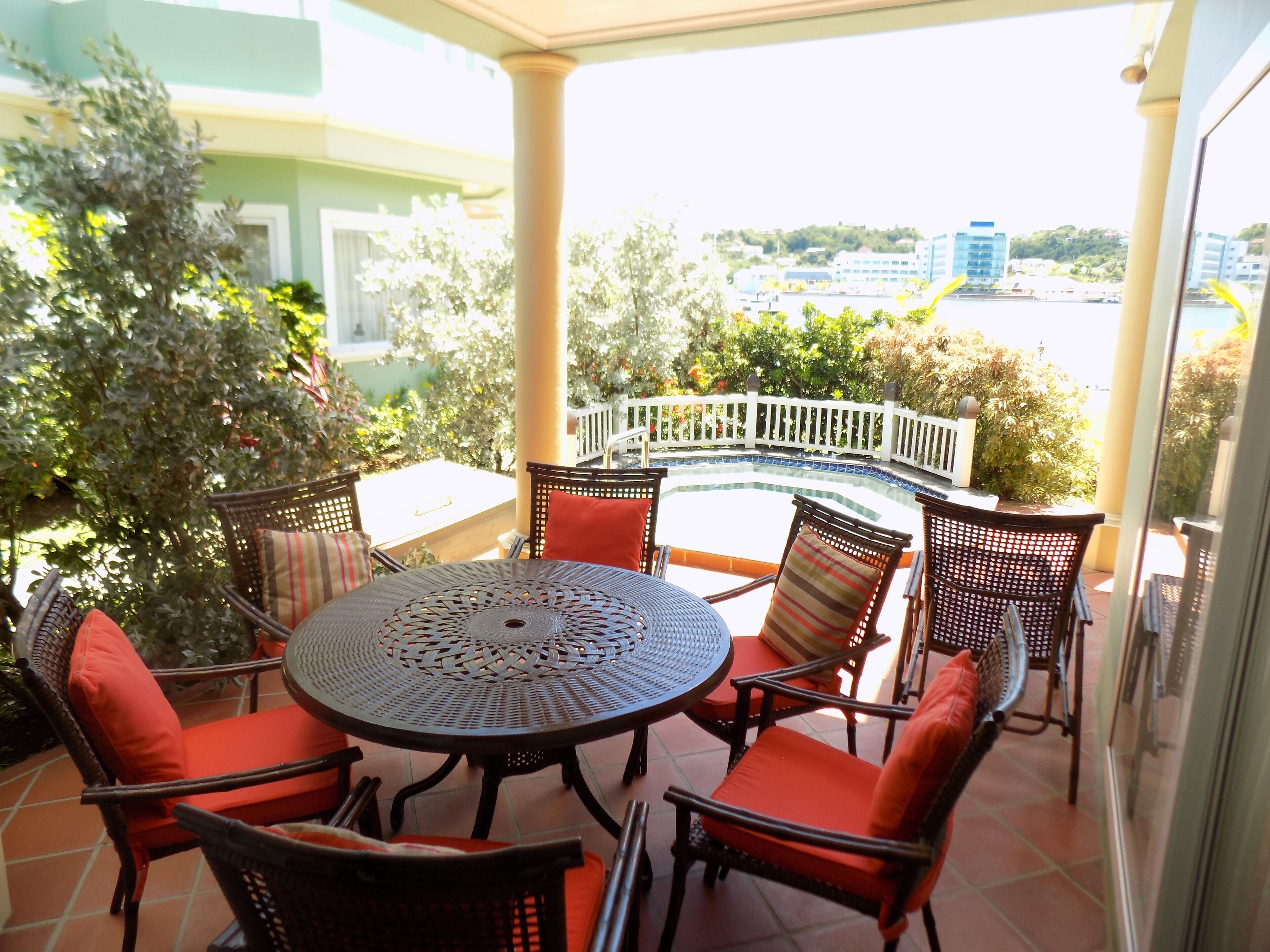 A covered outdoor patio with a round table and wicker chairs, surrounded by greenery and a view of buildings in the distance.