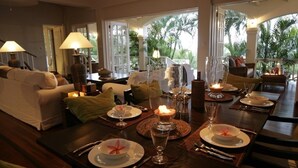 A dining area with a table set for four, surrounded by a sofa and chairs, with large windows offering a view of palm trees. - Beautiful Caribbean Style 2-bed Family Villa - Villa Kessi 2 Bedroom Villa by RedAwning (Gros Islet)