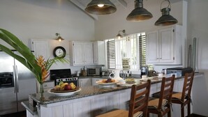 A kitchen with white cabinets, a central island with chairs, a clock on the wall, and a sign on the window. - Beautiful Caribbean Style 2-bed Family Villa - Villa Kessi 2 Bedroom Villa by RedAwning (Gros Islet)