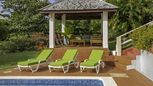 Ein Poolbereich mit drei grünen Liegestühlen, einer überdachten Sitzgelegenheit und einer Treppe, die in einen Garten führt.