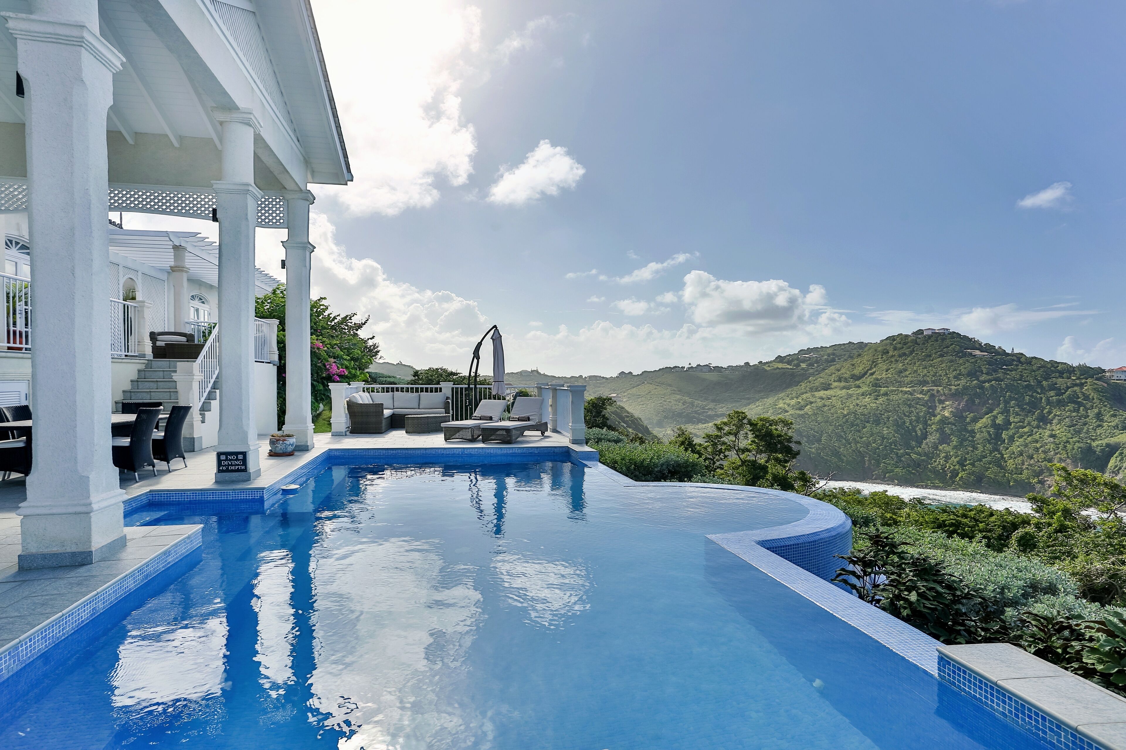 A pool with a view of a beach and mountains, surrounded by a covered patio with seating and a table.