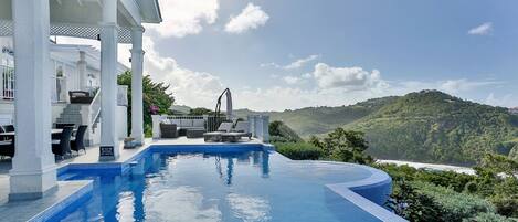 A pool with a view of a beach and mountains, surrounded by a covered patio with seating and a table.