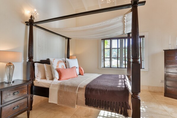 A four-poster bed with a canopy, a nightstand with a lamp, and a dresser in a well-lit bedroom with a window.
