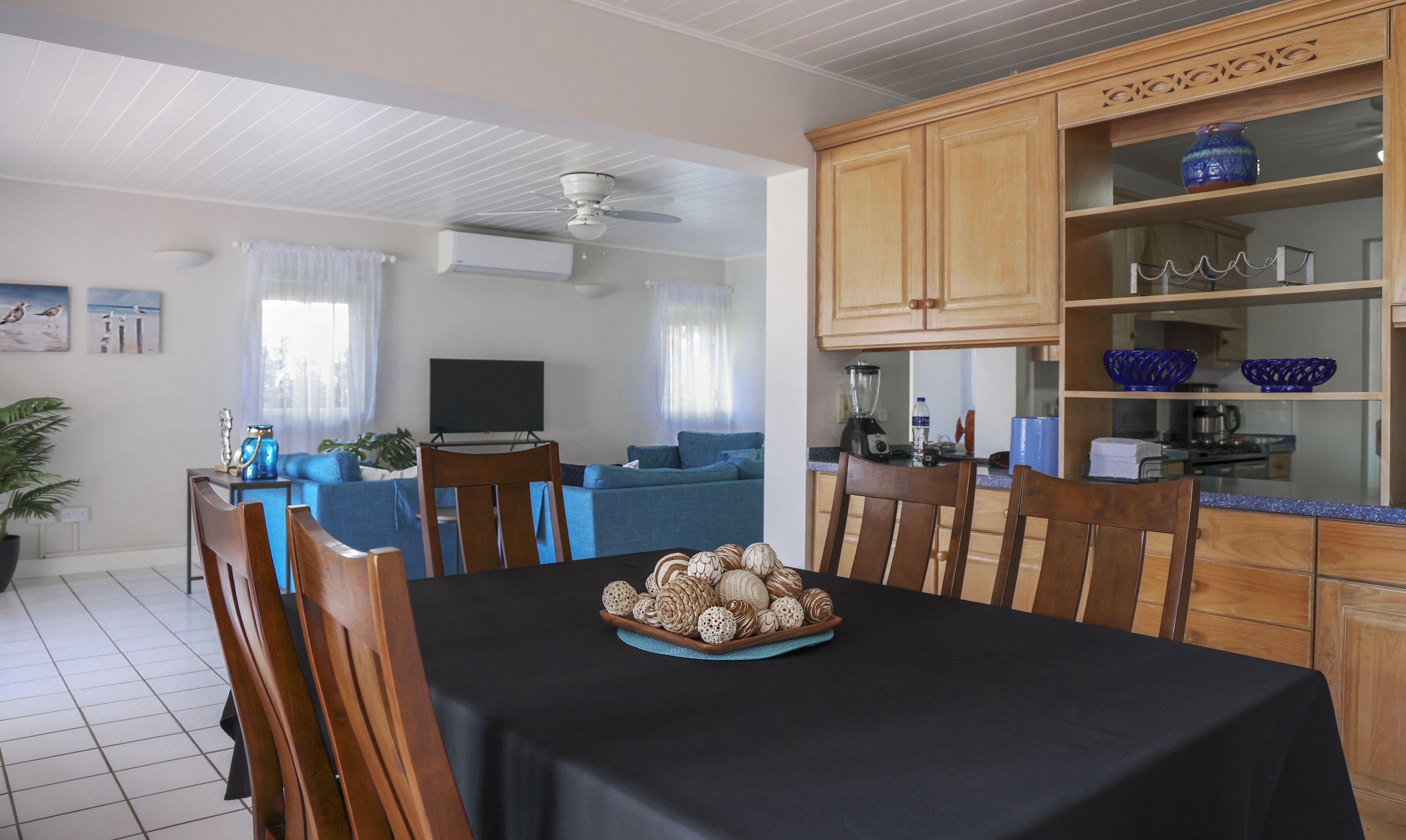 En matplats med ett bord för fyra personer, ett kök med träskåp och blå dekorativa detaljer samt ett vardagsrum med en soffa och en platt-TV.