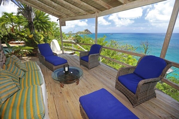A covered outdoor seating area with wicker chairs and cushions, overlooking a tropical beach and ocean.