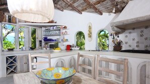 A kitchen with a wooden table, chairs, and a bowl. There are shelves with dishes and a window with a view of greenery. - Smuggler's Nest - Exotic & Romantic Villa 2 Bedroom Villa by RedAwning (Gros Islet)