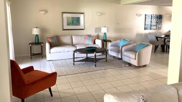 A living room with a white sofa, a teal armchair, and a red chair. There are two side tables with lamps, a ceiling fan, and a framed picture on the wall.