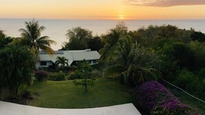 A house with a view of the sunset over the ocean, surrounded by lush greenery and palm trees.