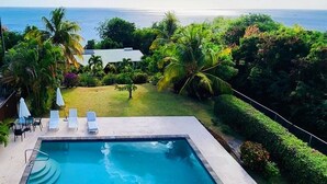 A swimming pool with lounge chairs and umbrellas, surrounded by lush greenery and a view of the ocean.