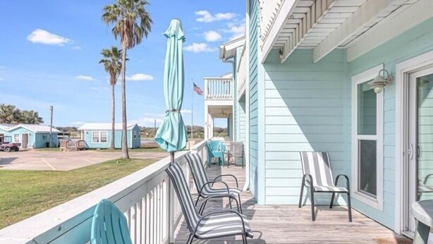 A wooden deck with chairs and a covered area, overlooking a beach with palm trees and a clear sky. - Sunset Breeze 3 Bedroom Condo by RedAwning (Rockport)