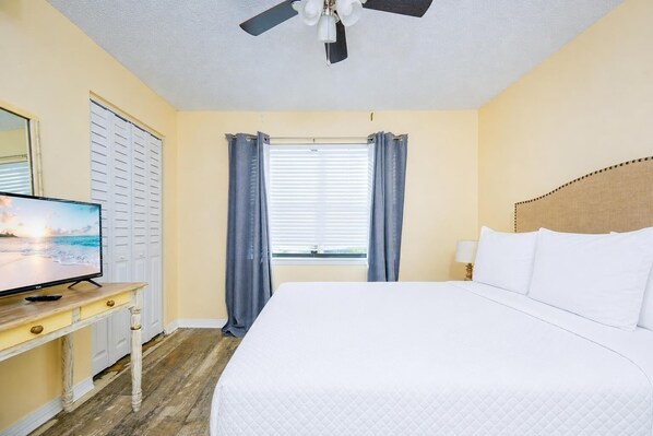 Quarto de hotel com cama, televisão sobre uma cômoda de madeira, ventilador de teto e janela com cortinas.