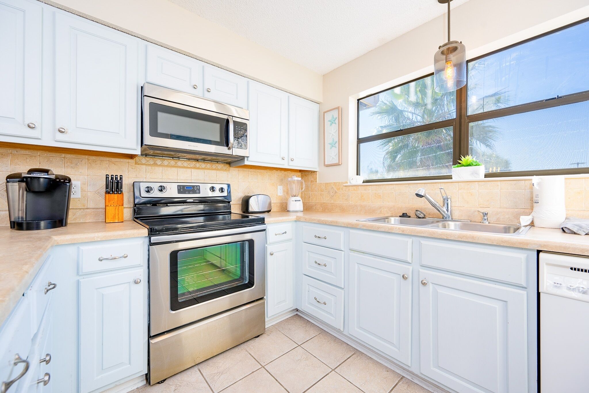 A kitchen with white cabinets, a stove, a microwave, a sink, and a window with a view of palm trees.