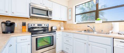 A kitchen with white cabinets, a stove, a microwave, a sink, and a window with a view of palm trees.