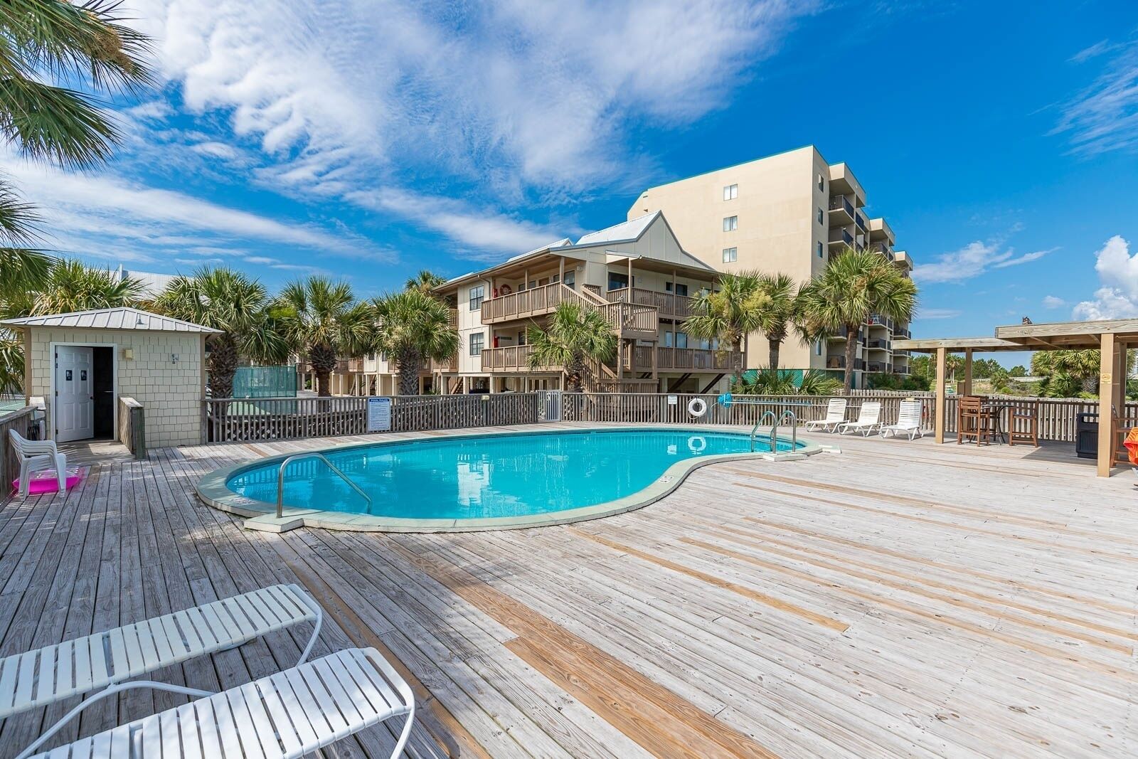 A pool area with a wooden deck, lounge chairs, and a building with balconies.