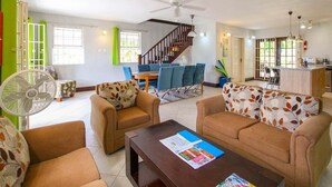A spacious living room with a sofa set, a coffee table, and a fan. There is a staircase leading to another level, a dining area, and a kitchen visible in the background.