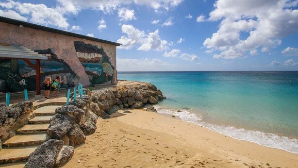 A beach with a building featuring a mural, a staircase leading to the building, and a clear view of the ocean.
