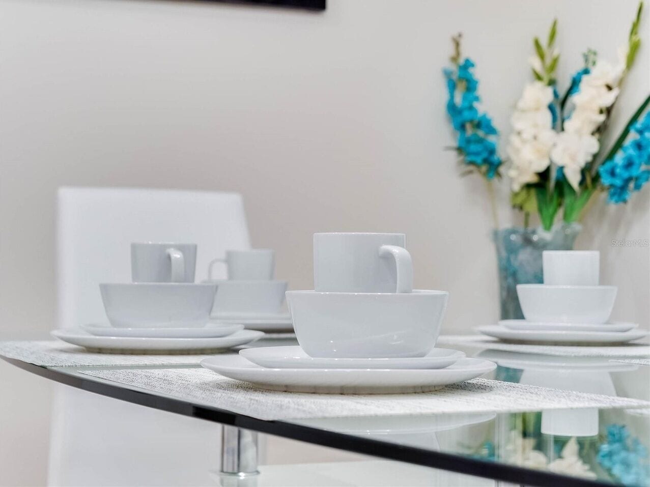 A table set with white cups and saucers, a vase with flowers, and a framed picture on the wall.