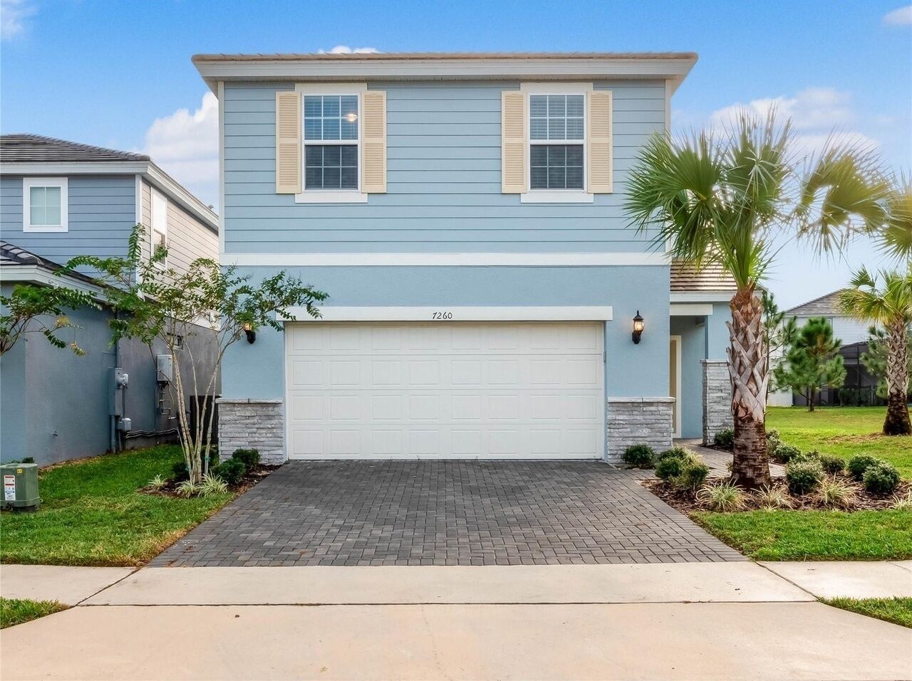 A two-story house with a garage, a paved driveway, and a well-maintained lawn with palm trees.