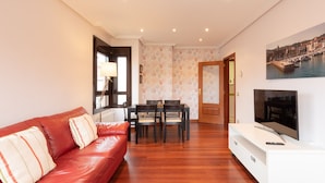 Una sala de estar con un sofá de cuero rojo, una mesa de comedor con sillas, un televisor sobre un mueble blanco y un cuadro de un paisaje urbano en la pared.