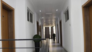 A long, narrow hallway with wooden doors on either side, a central potted plant, and a metal railing in the foreground. - James Court Hotel & Luxury Apartments (Lekki)