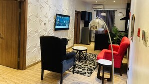 A modern living room with a black armchair, a red sofa, a small round table, and a TV mounted on the wall. - James Court Hotel & Luxury Apartments (Lekki)