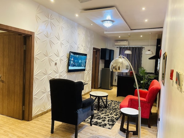 A modern living room with a black armchair, a red sofa, a small round table, and a TV mounted on the wall. - James Court Hotel & Luxury Apartments (Lekki)