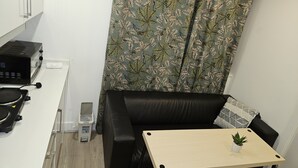 A compact living space with a kitchenette, a black leather sofa, a small wooden table with a plant, and a curtain with a leaf pattern. - 1 Bed Flat - Sleeps 2 - Parking - Garden (London)