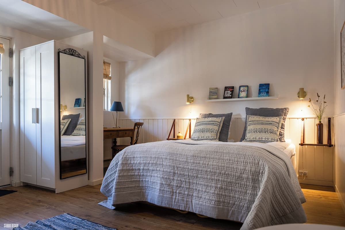 Quarto com uma cama grande, mesa de cabeceira, cadeira, escrivaninha, espelho e prateleira com livros.